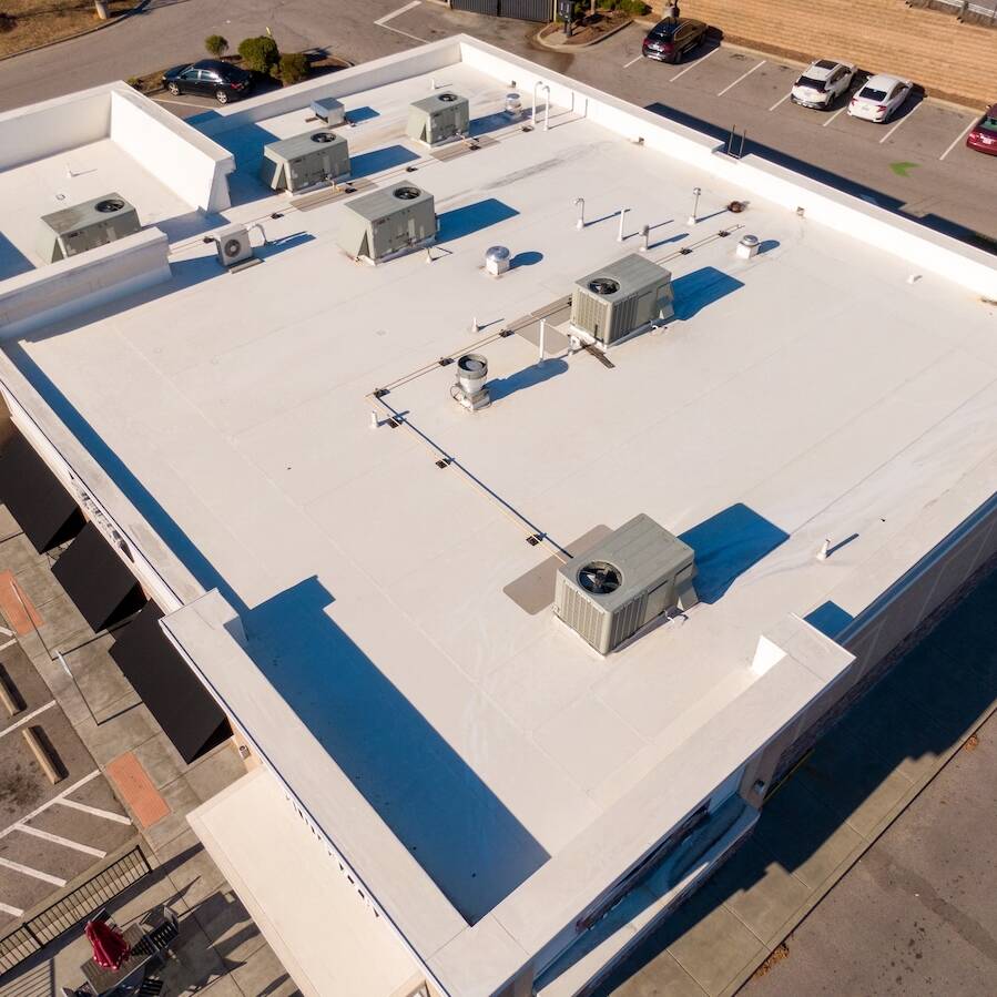 Aerial view of a flat, white commercial building rooftop with multiple HVAC units and pipes. Surrounding the building is a parking lot with cars and marked spaces, including accessible parking spots and green painted arrows.
