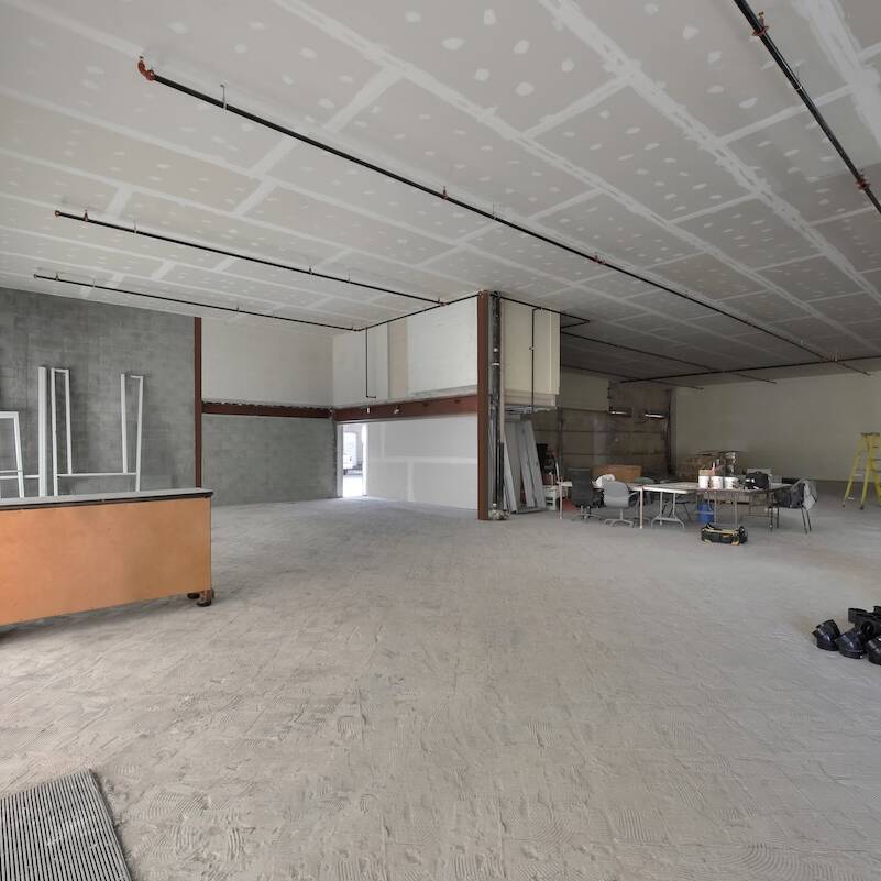 A large, unfinished commercial interior with exposed drywall, scattered construction materials, tables, chairs, a ladder, and boxes—ideal for renovation or water damage restoration. Large windows let in natural light from the left side.